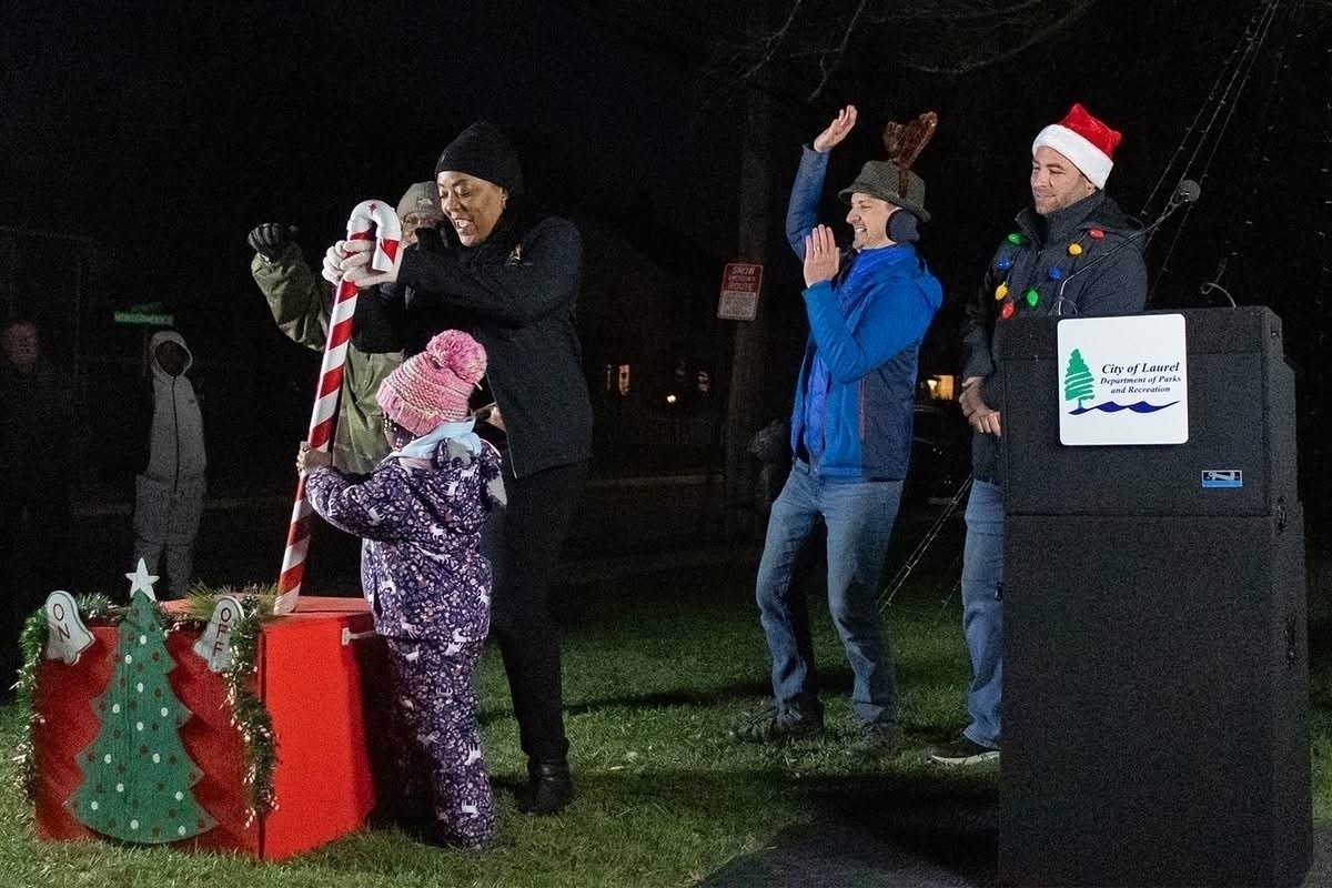 Auto-generated description: A festive outdoor event features people gathered around a candy cane decoration, with some wearing winter clothing and Santa hats.