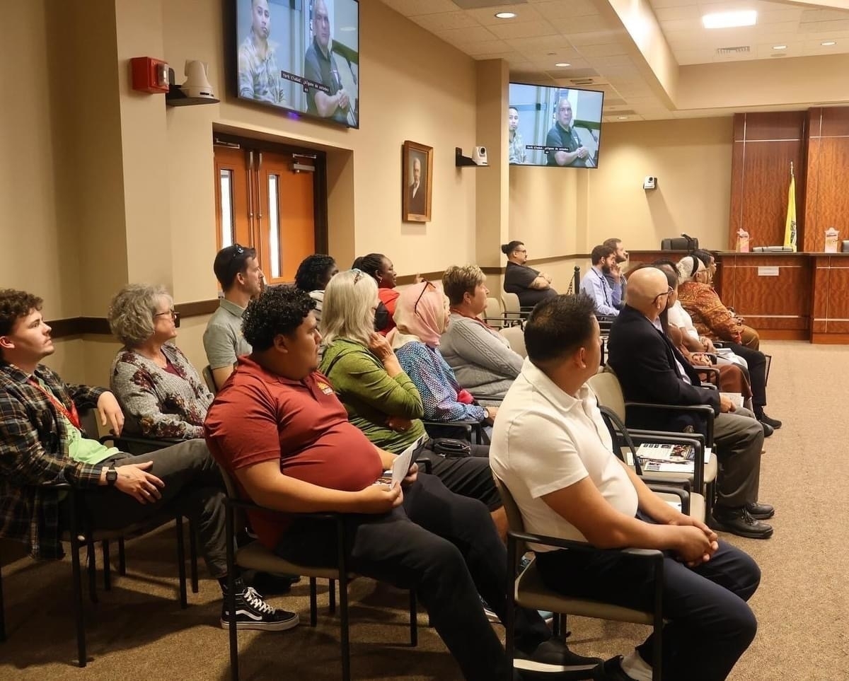 Auto-generated description: A group of people seated in a room with a meeting or presentation taking place, as shown on the mounted screens.