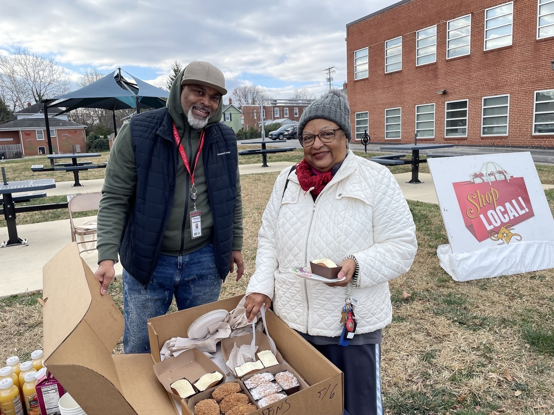 Auto-generated description: Two people are standing outdoors with a box of pastries and a Shop Local sign nearby.