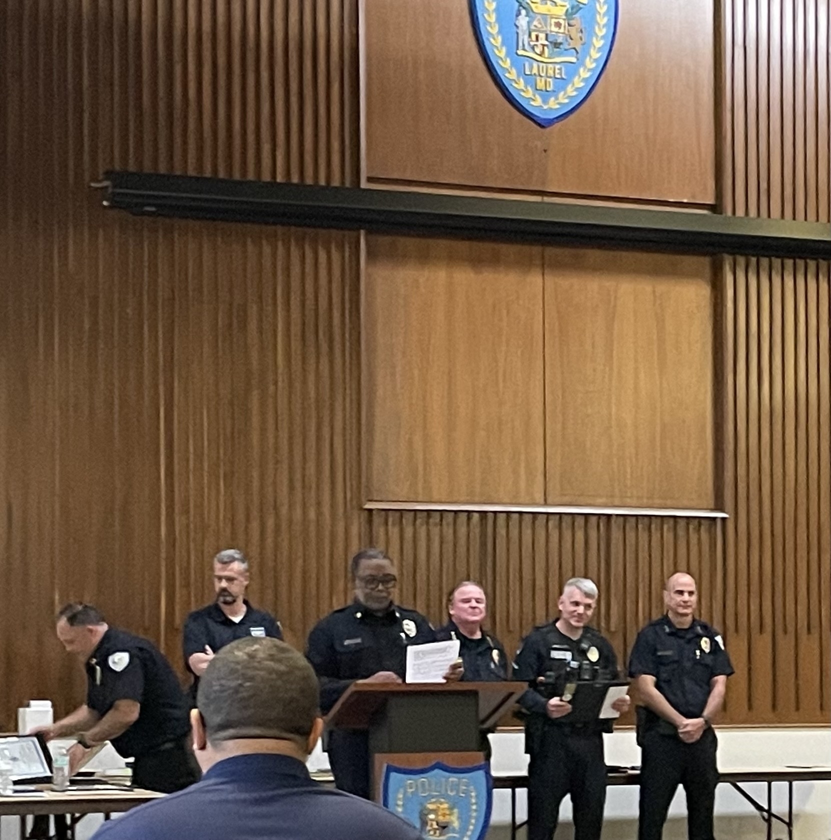 Auto-generated description: Several individuals wearing police uniforms are standing together inside a wood-paneled room with a police emblem on the wall.