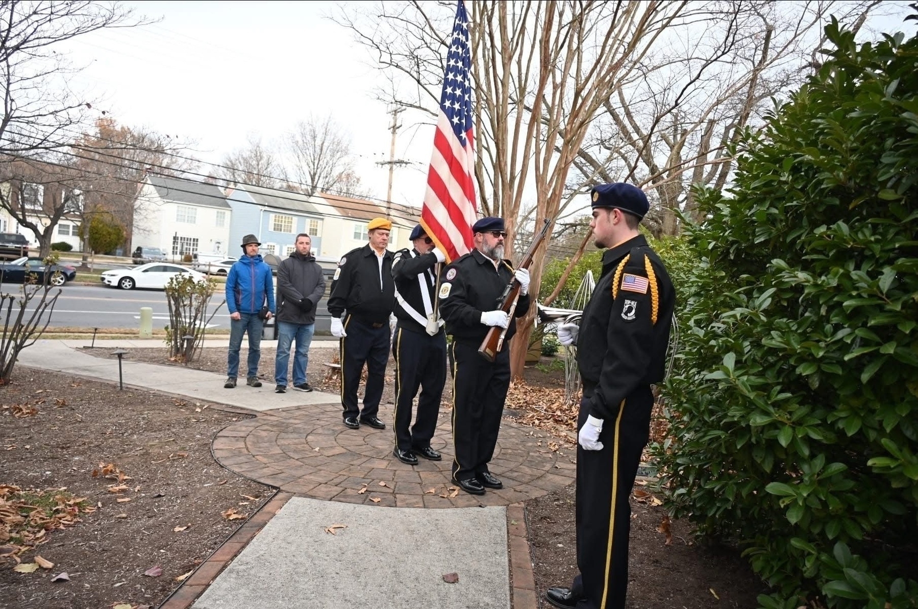 Auto-generated description: A group of people stand outside near a sidewalk, with some individuals in uniform holding an American flag, while others watch nearby.