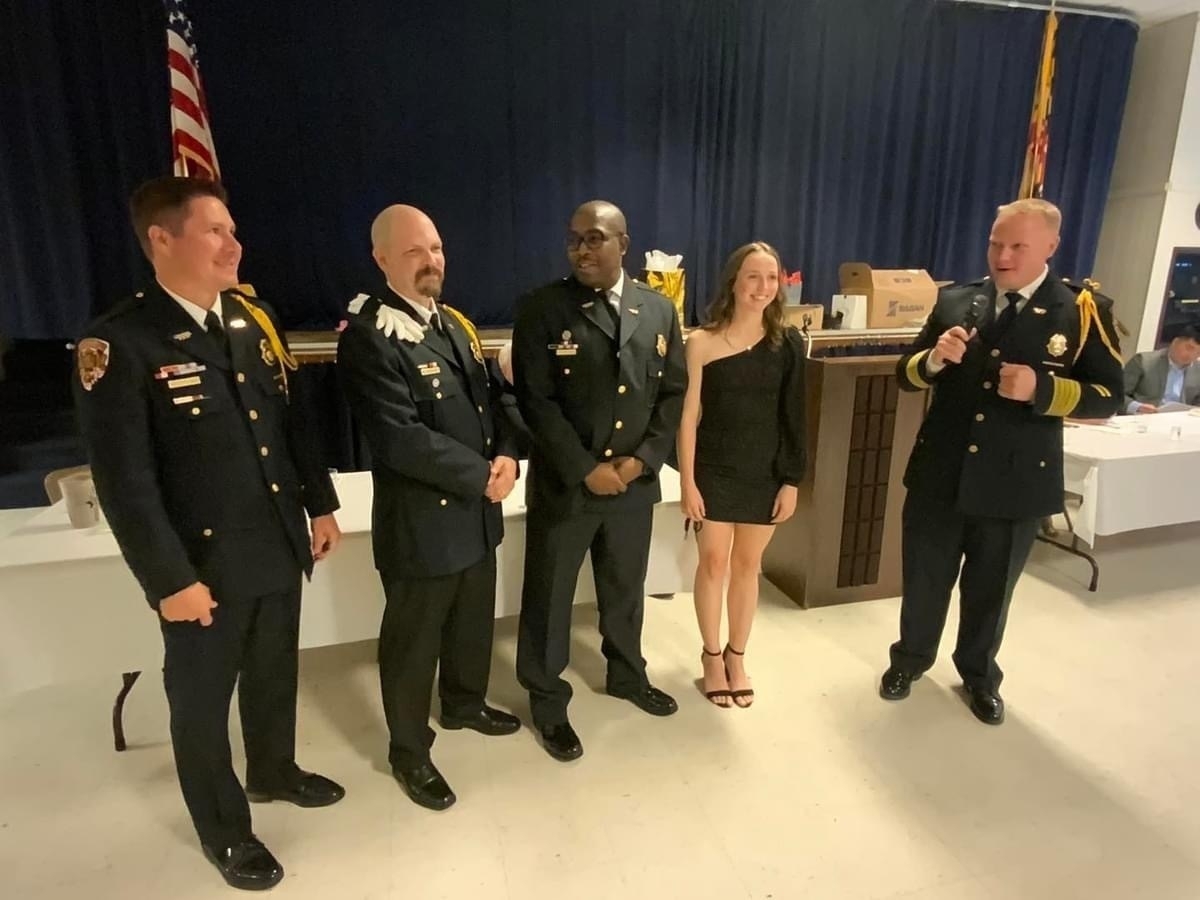 Auto-generated description: Five people are standing together in a room, with some wearing formal uniforms and one in a black dress, in front of a table with an American flag in the background.