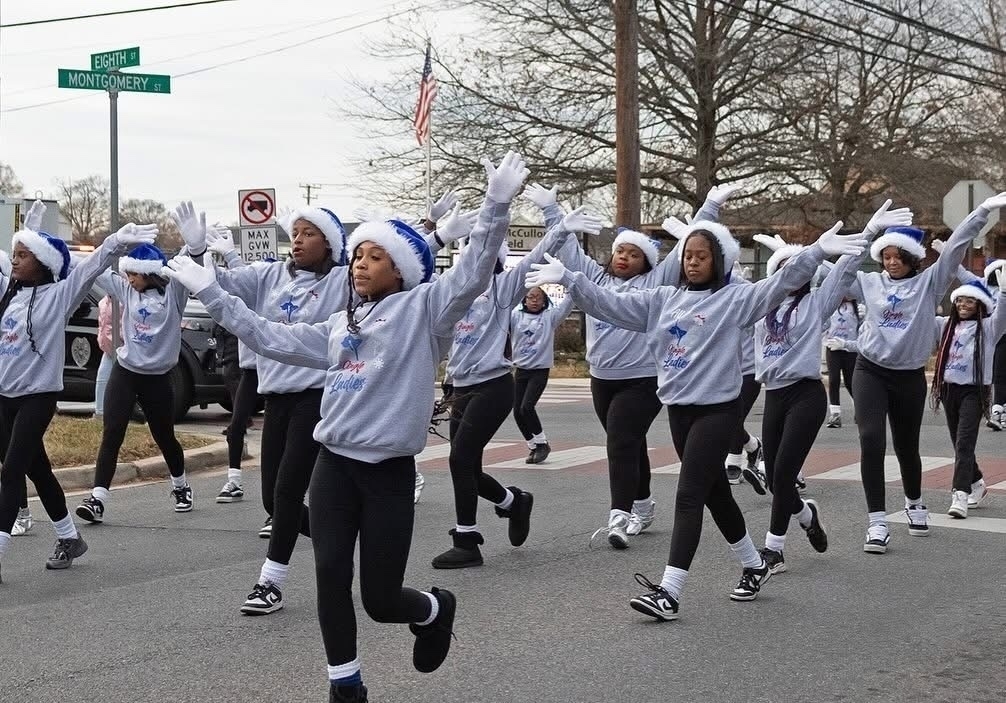 Auto-generated description: A group of people wearing matching outfits and Santa hats are marching together, performing a synchronized routine on a street.