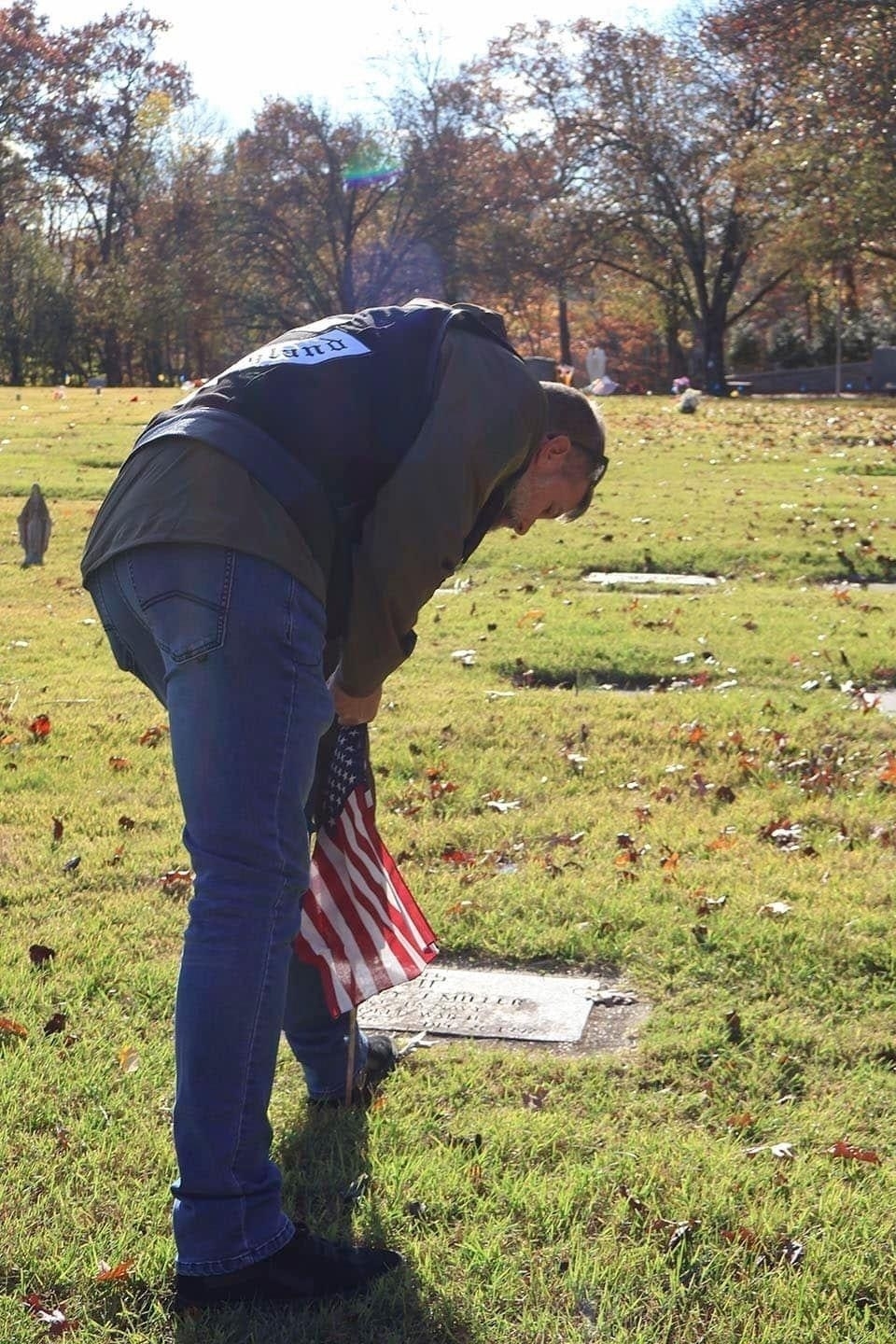 Auto-generated description: A person is bending over a grave marker while holding an American flag in a cemetery surrounded by autumn trees.