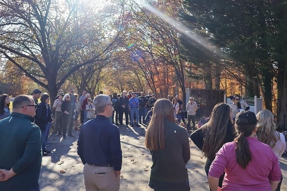 Auto-generated description: A group of people is gathered outdoors, listening to someone speak, surrounded by trees with autumn foliage.