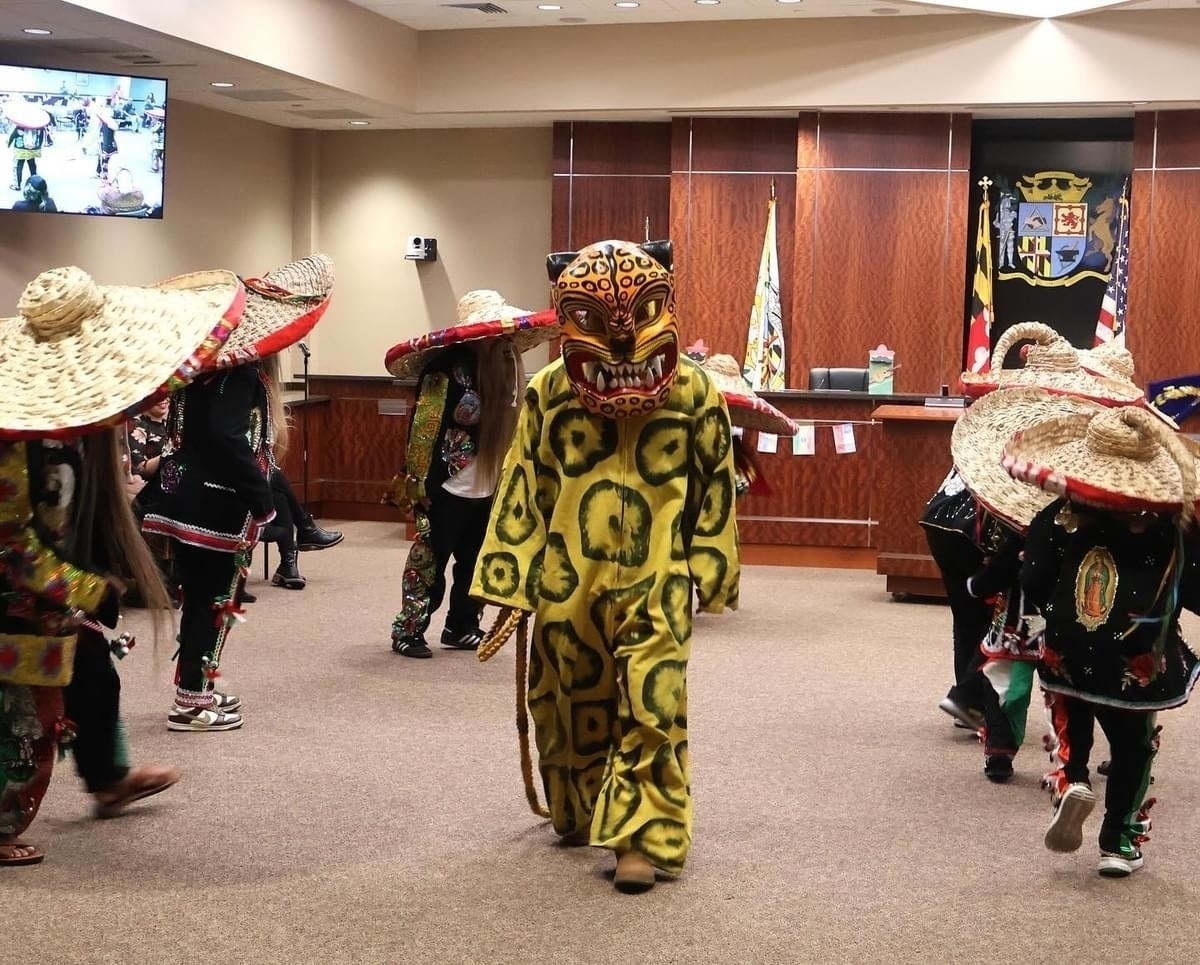 Auto-generated description: A group of people wearing colorful costumes and straw hats are performing in a room with wood paneling and a seal on the wall.