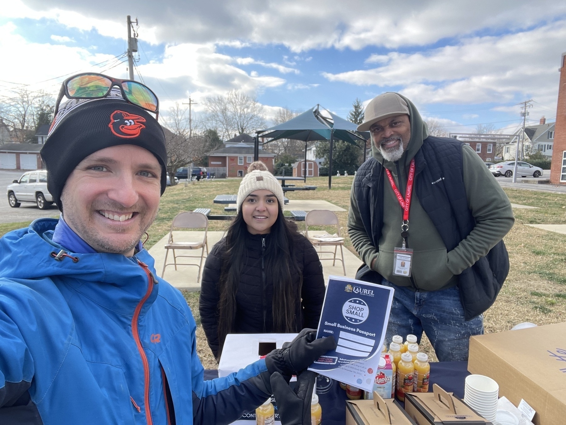 Auto-generated description: Three people are posing for a selfie at an outdoor table with drinks and a plaque.