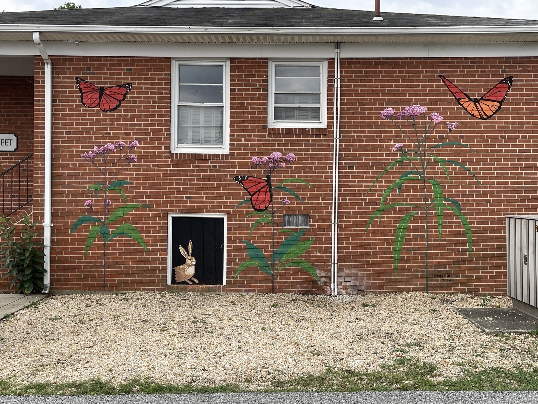 Auto-generated description: A brick wall features murals of monarch butterflies and tall, flowering plants, with a door where a small rabbit statue peeks out.