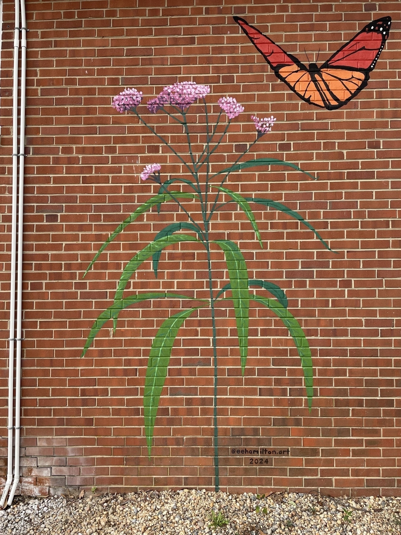 Auto-generated description: A brick wall features a mural of tall, green plants with pink flowers and an orange and black butterfly.