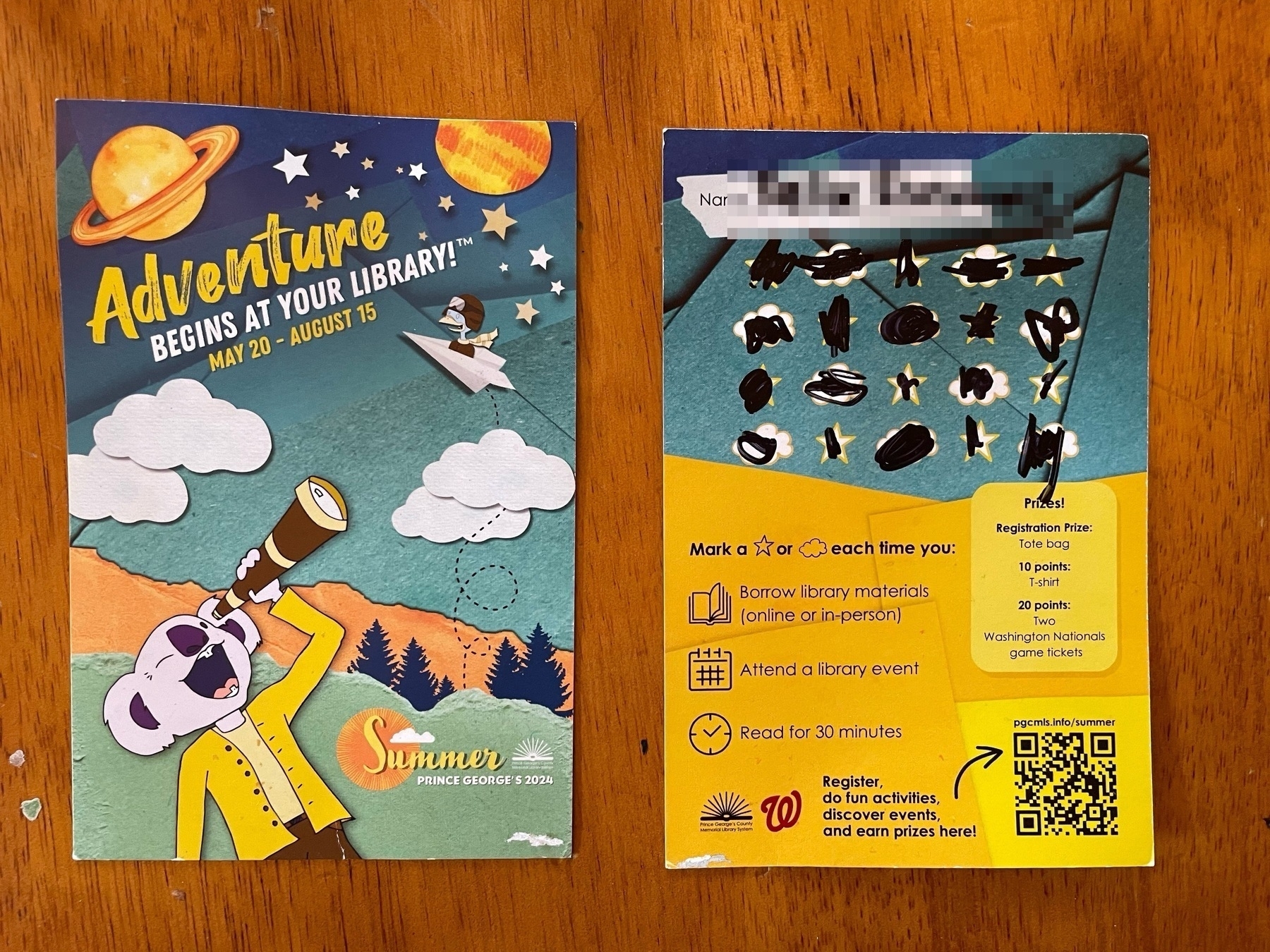 Auto-generated description: A promotional poster and a completed library event card with stamps featuring an outer space theme and summer reading incentive details are displayed on a wooden surface.