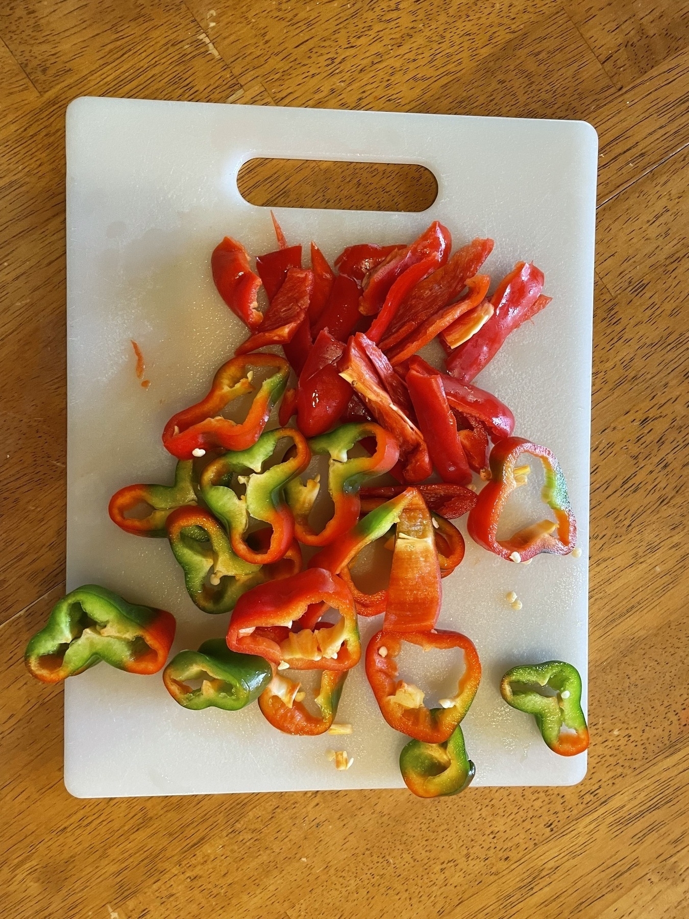 Auto-generated description: Sliced red and green bell peppers are arranged on a white cutting board placed on a wooden table.