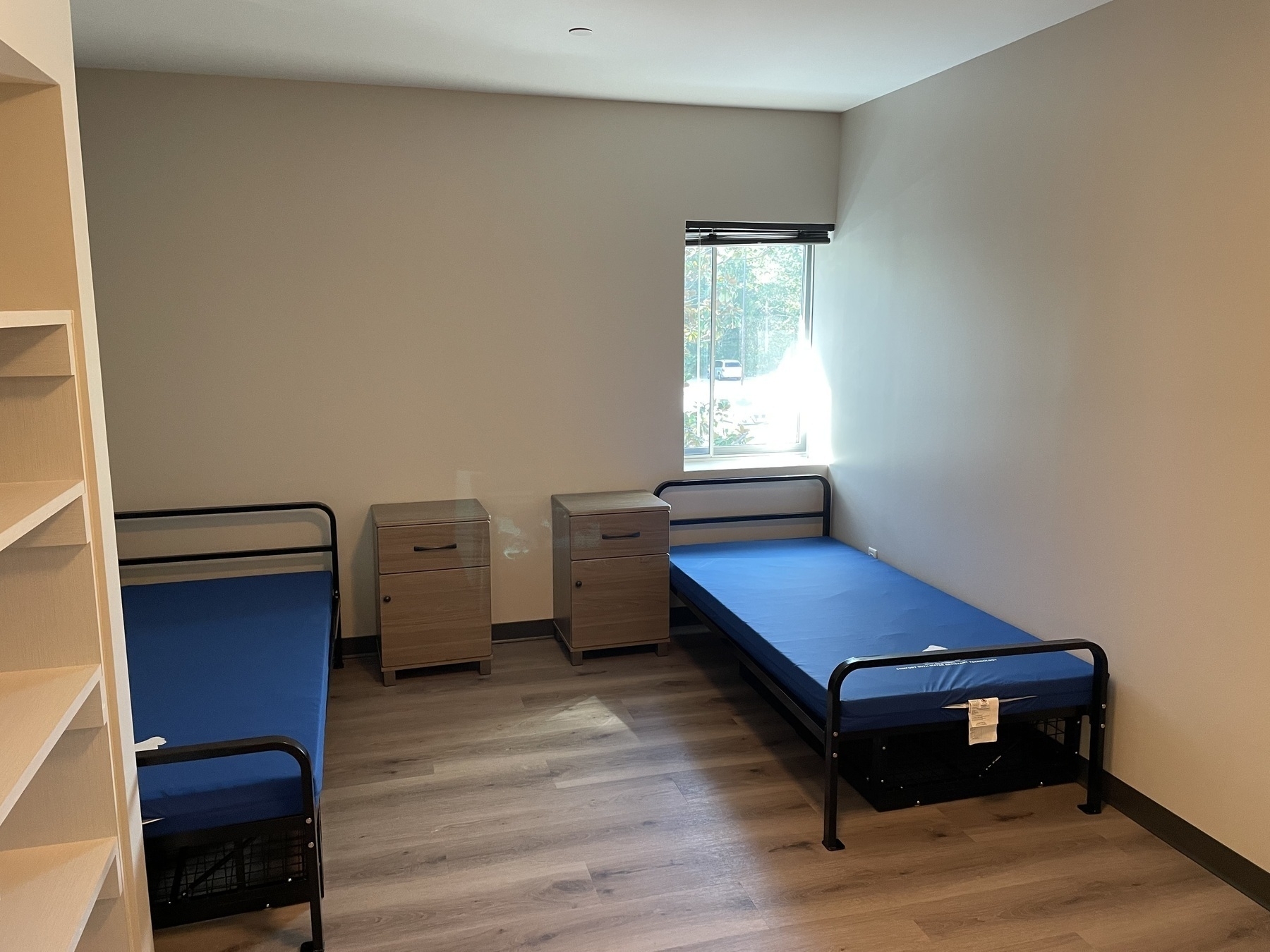 Auto-generated description: Two single beds with blue mattresses are set against opposite walls in a simple, sparsely furnished room with a window.