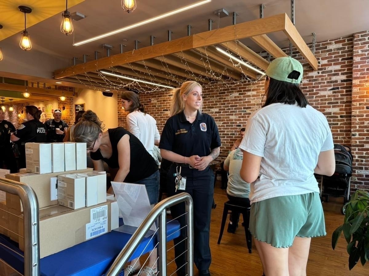 Auto-generated description: A group of people are interacting inside a café or similar establishment, with boxes on a cart and a person wearing a firefighter shirt.