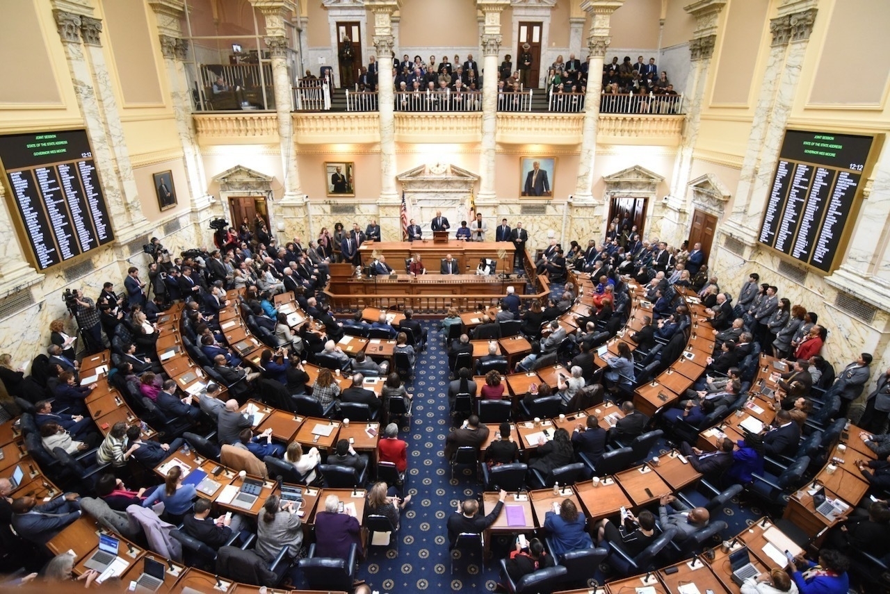 Auto-generated description: A large assembly is gathered in a government chamber with people seated in desks and a group at the front podium.