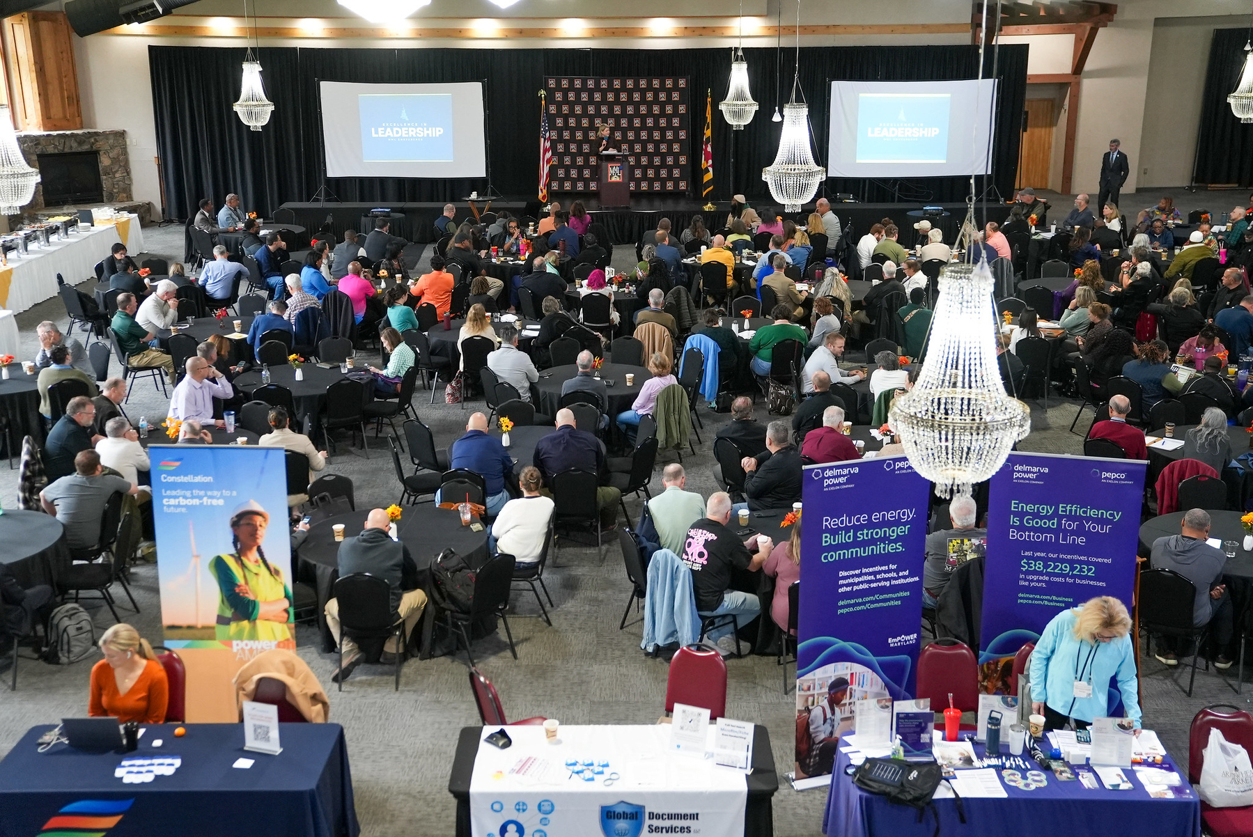 Auto-generated description: A large conference hall is filled with attendees seated at tables, listening to a presentation with promotional banners visible in the foreground.