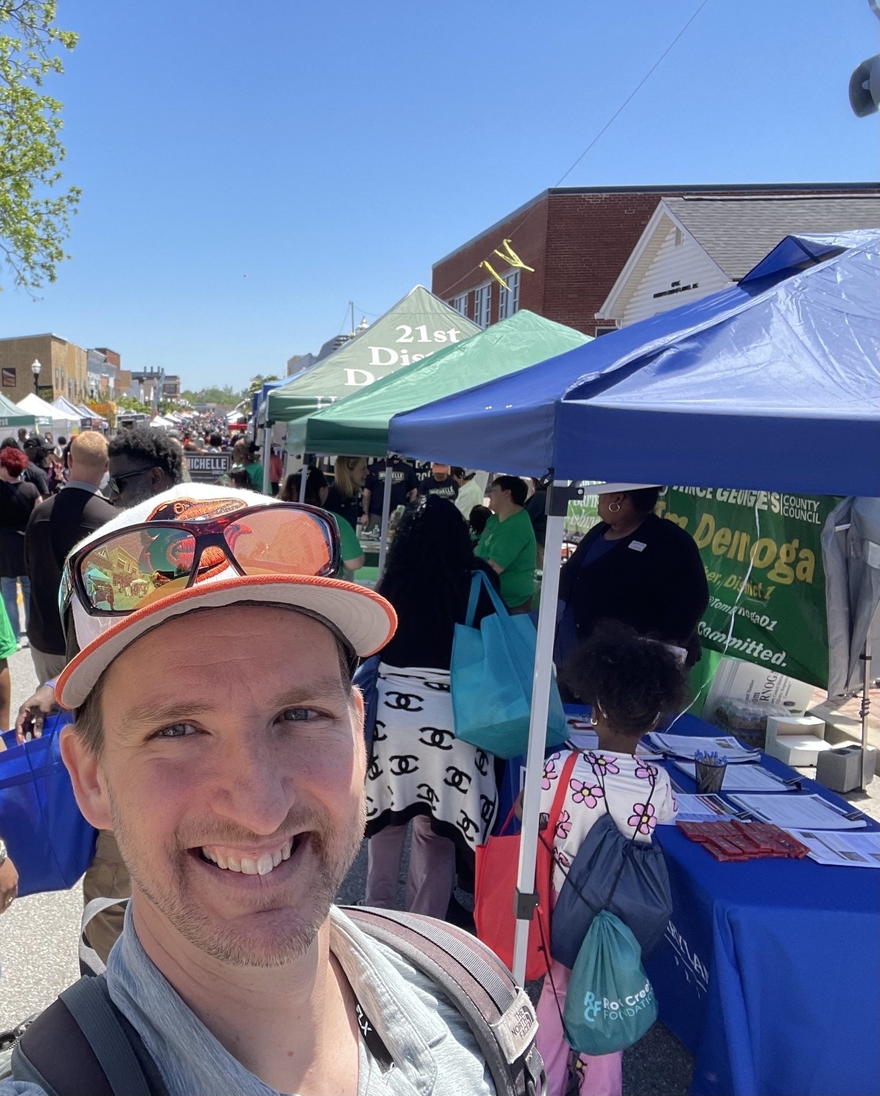 Auto-generated description: A man taking a selfie at an outdoor event with tents, booths, and people in the background.