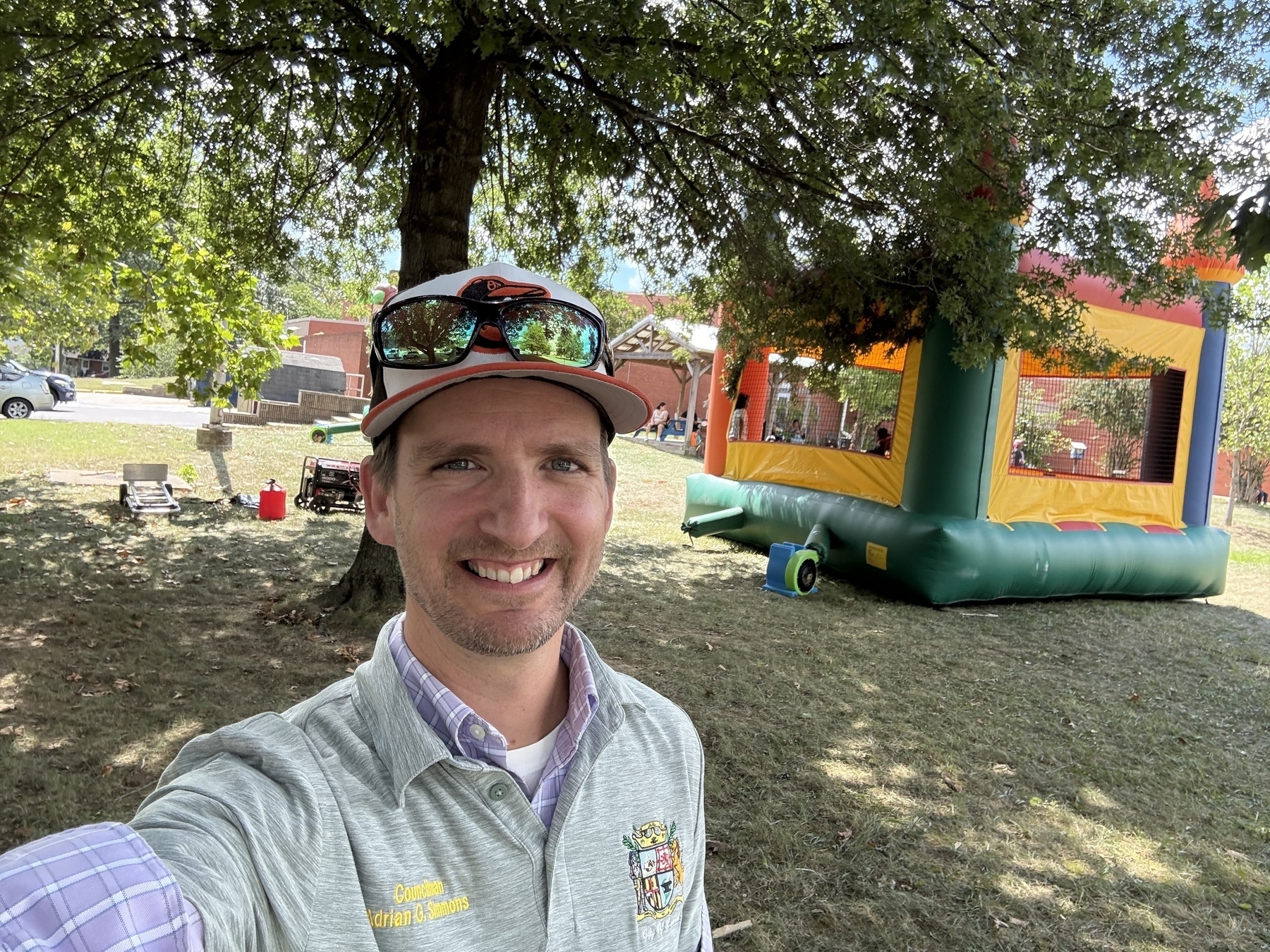Auto-generated description: A man wearing a cap and sunglasses is smiling in a park with a colorful bounce house in the background.