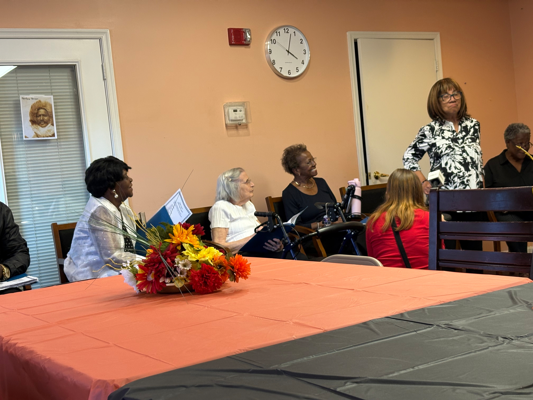 Auto-generated description: A group of people is gathered around tables with fall-themed decorations, while a woman stands and appears to be speaking.