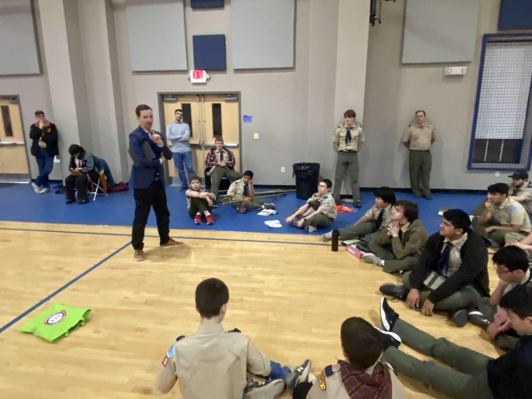 Auto-generated description: A person is speaking to a group of scouts seated on the floor in a gymnasium.