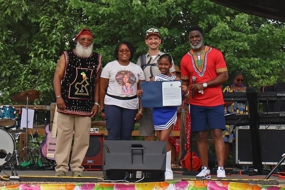Auto-generated description: A group of people stands together outdoors, with one young woman holding an award, surrounded by musical instruments and vibrant decorations.