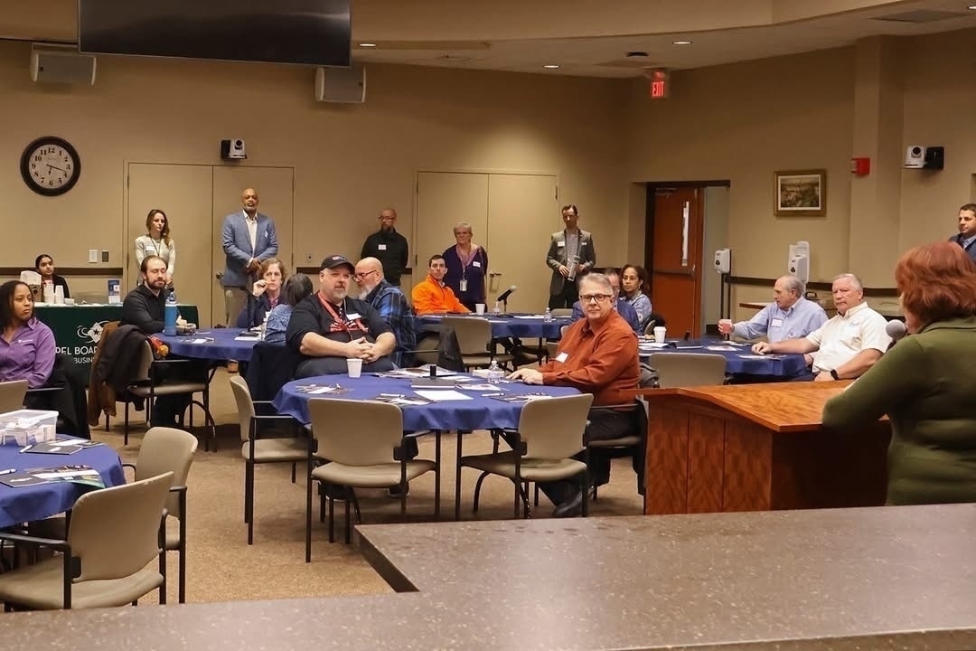 Auto-generated description: People are seated and standing around tables in a conference or meeting room, listening to a speaker at a podium.