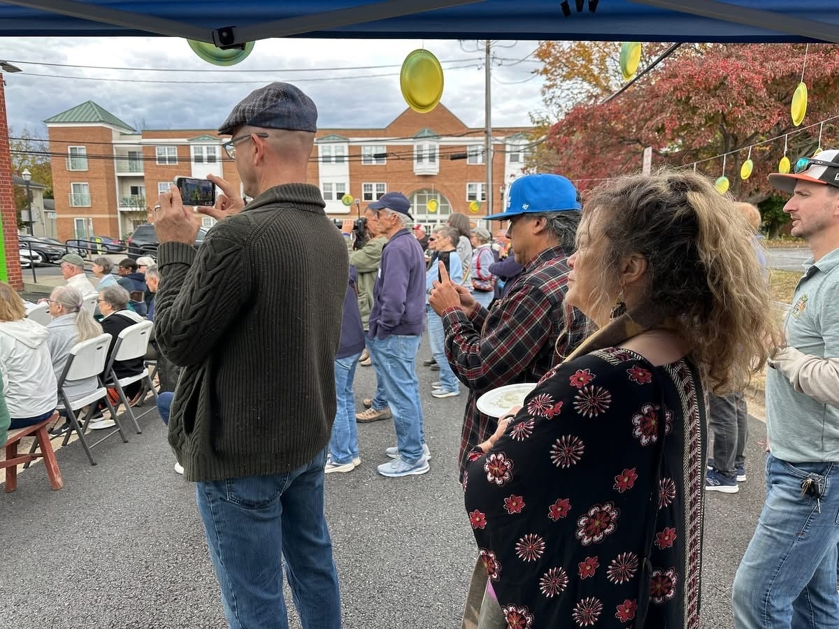 Auto-generated description: People are standing and seated at an outdoor event, some taking photos with their phones.