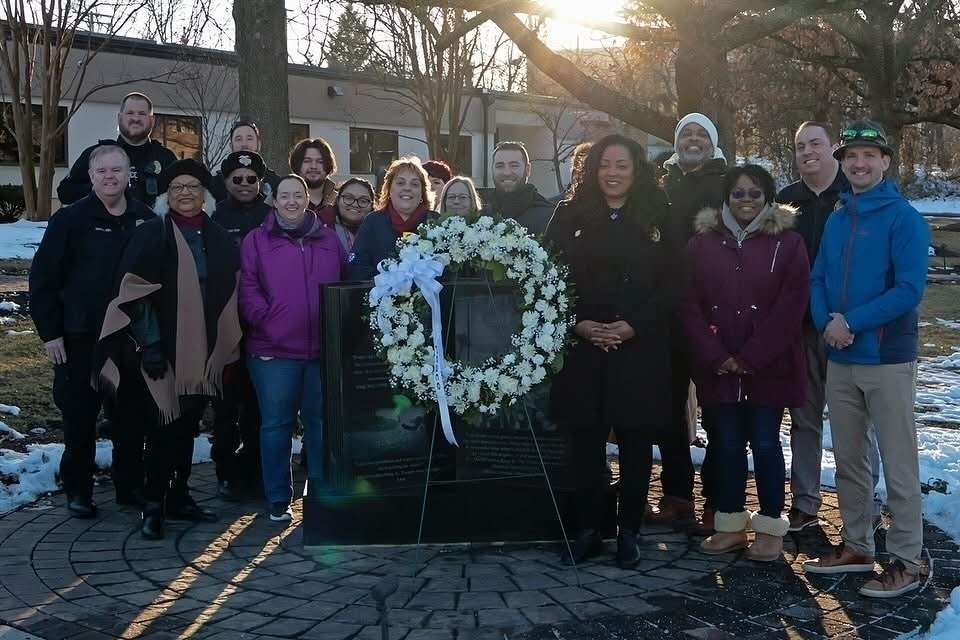 Auto-generated description: A group of people is gathered outdoors around a memorial plaque with a wreath in a snowy setting.