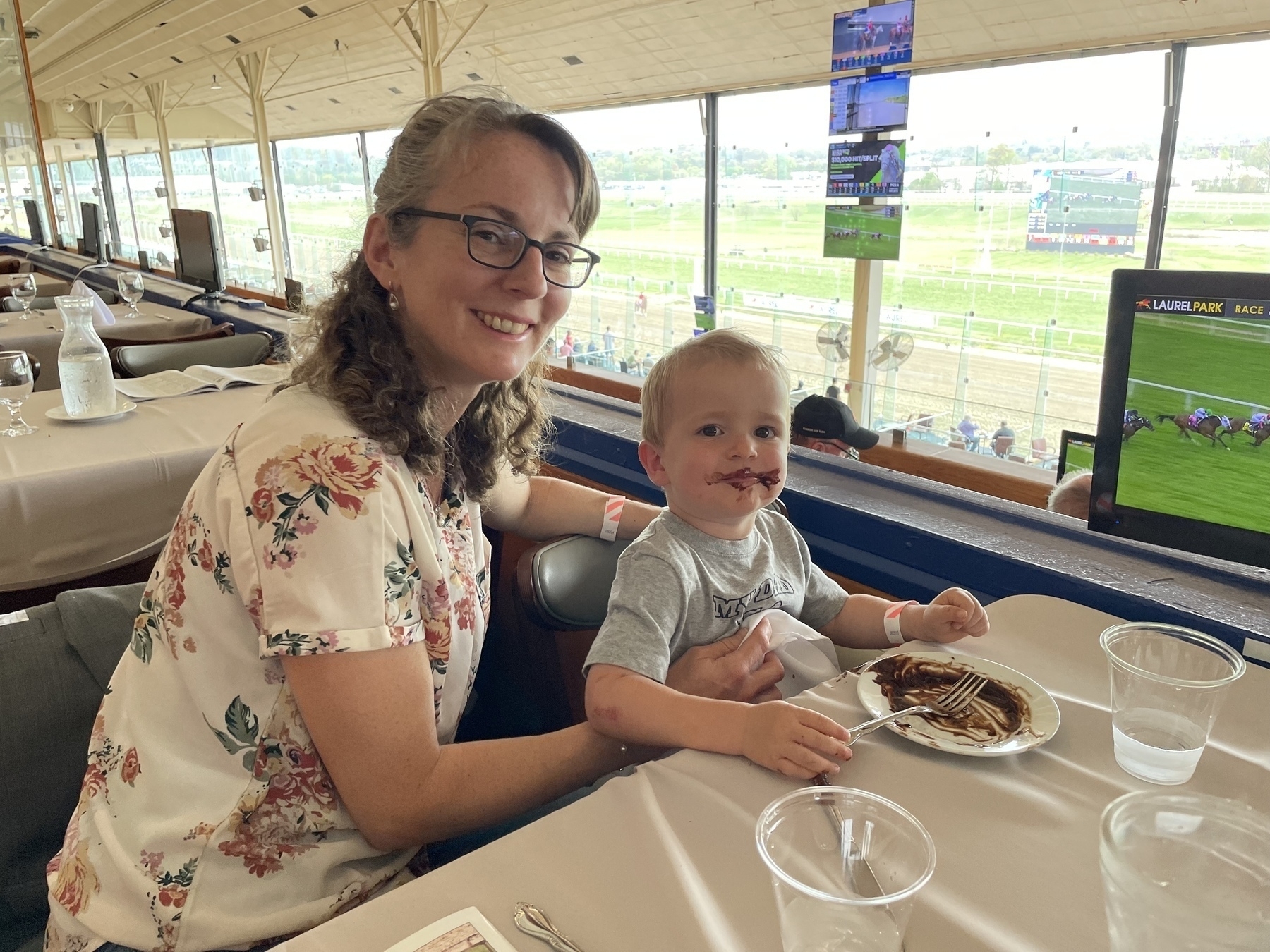 Auto-generated description: A woman with glasses and a young child, who has chocolate on their face, are sitting at a table with plates and watching horse racing at a track.