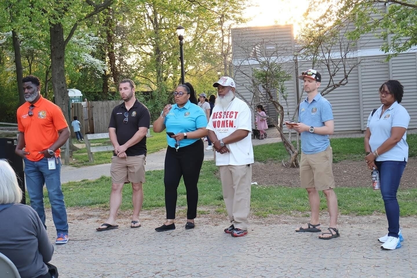 Auto-generated description: Six people standing outdoors in a line, facing the camera, with trees and a building in the background.