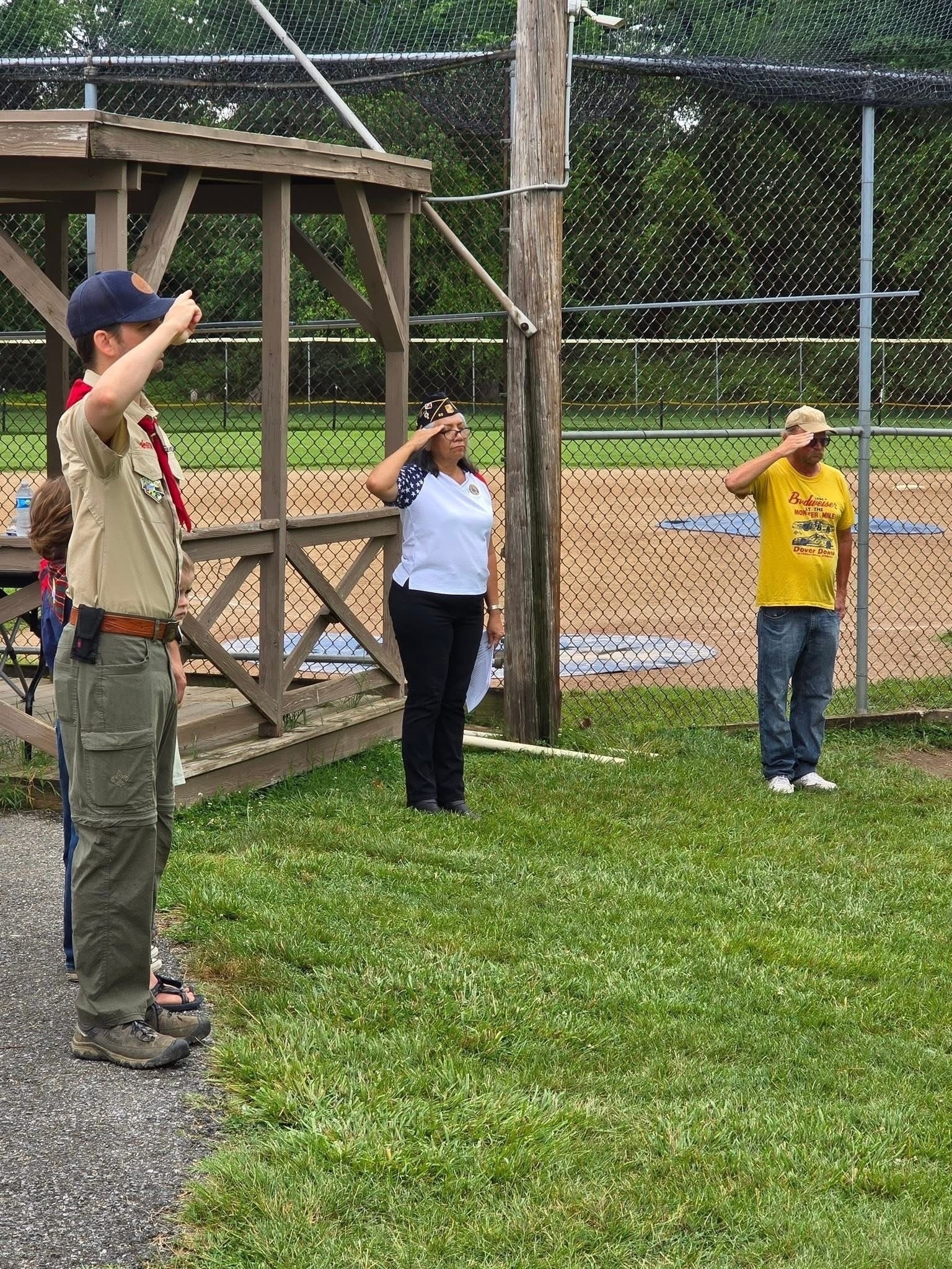 Auto-generated description: People are standing outdoors near a baseball field, saluting with their right hands raised.
