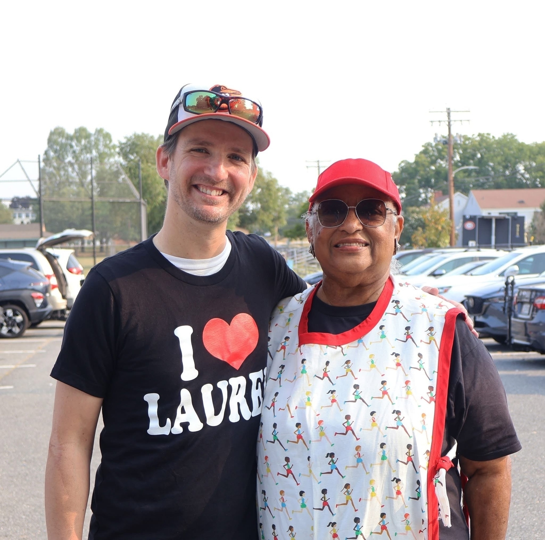 Auto-generated description: Two people are smiling outdoors in a parking lot, one wearing a cap and I ♥ LAUREL shirt, the other in glasses and a red cap.