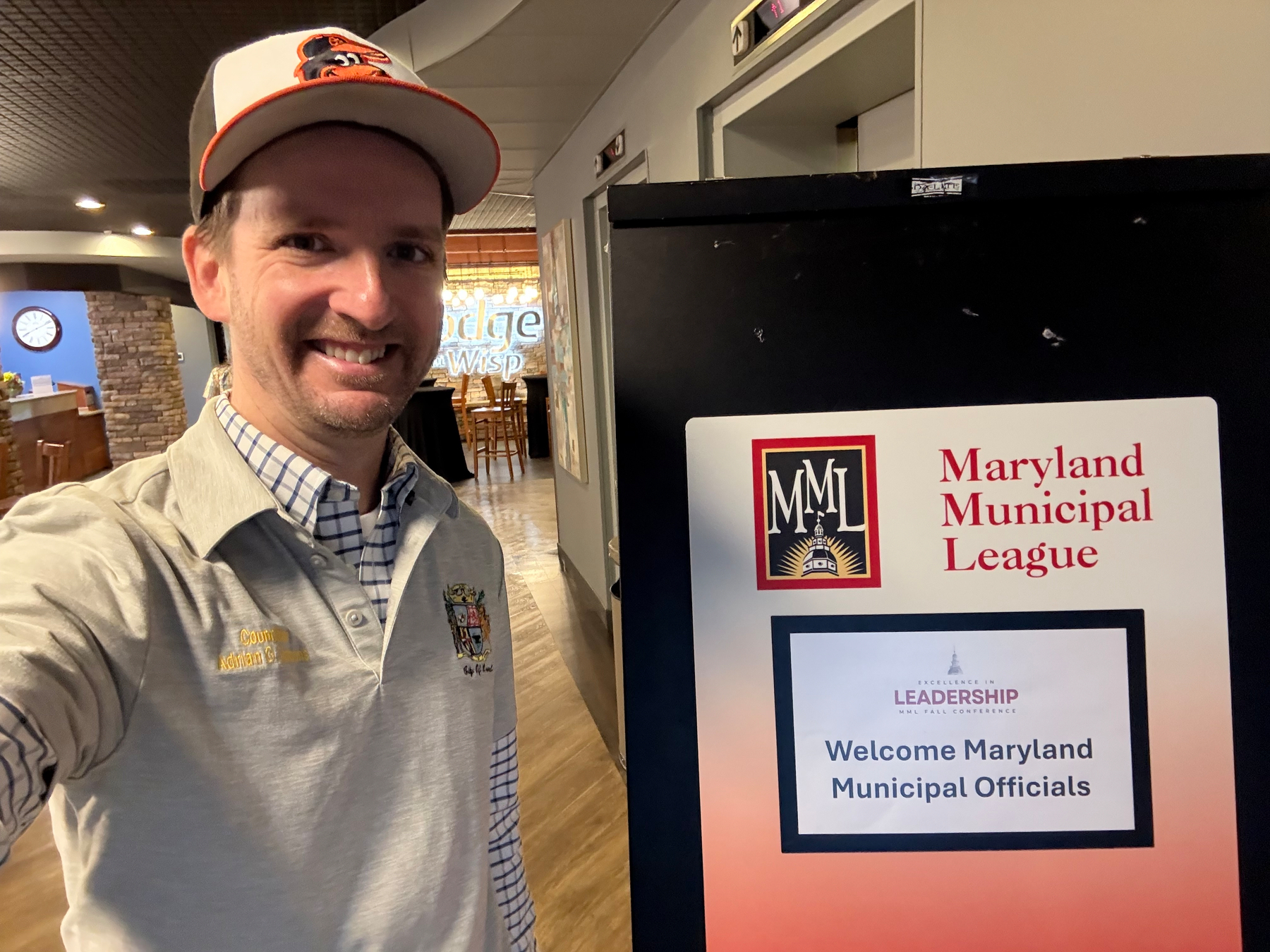 Auto-generated description: A smiling man wearing a cap takes a selfie next to a sign for a Maryland Municipal League leadership event.