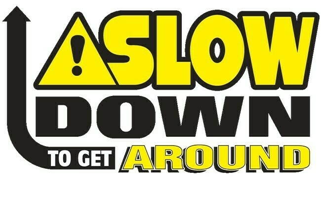 Auto-generated description: A bold yellow and black road safety sign with an exclamation mark, urging drivers to Slow Down to Get Around.