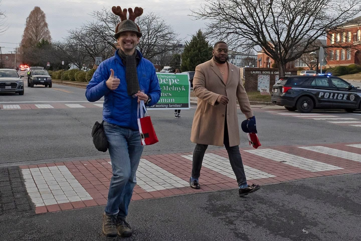 Auto-generated description: Two people are walking across a street, one wearing reindeer antlers and giving a thumbs-up while carrying a small red gift bag.