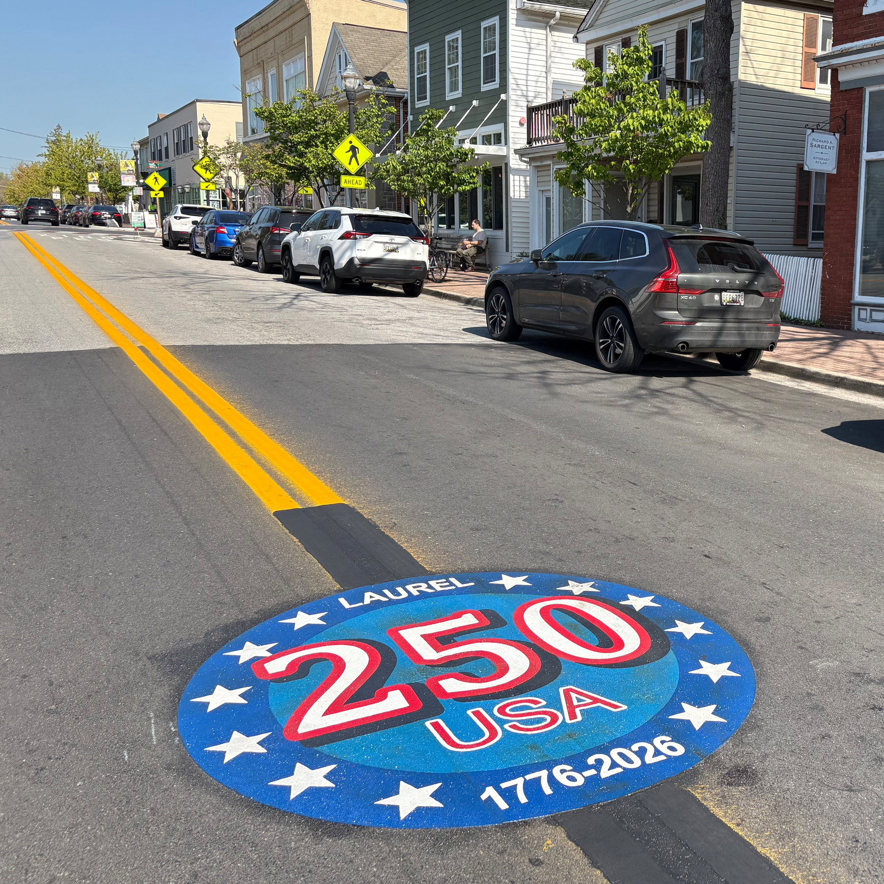 Auto-generated description: A 250th anniversary emblem is painted on a street with parked cars and buildings on either side.