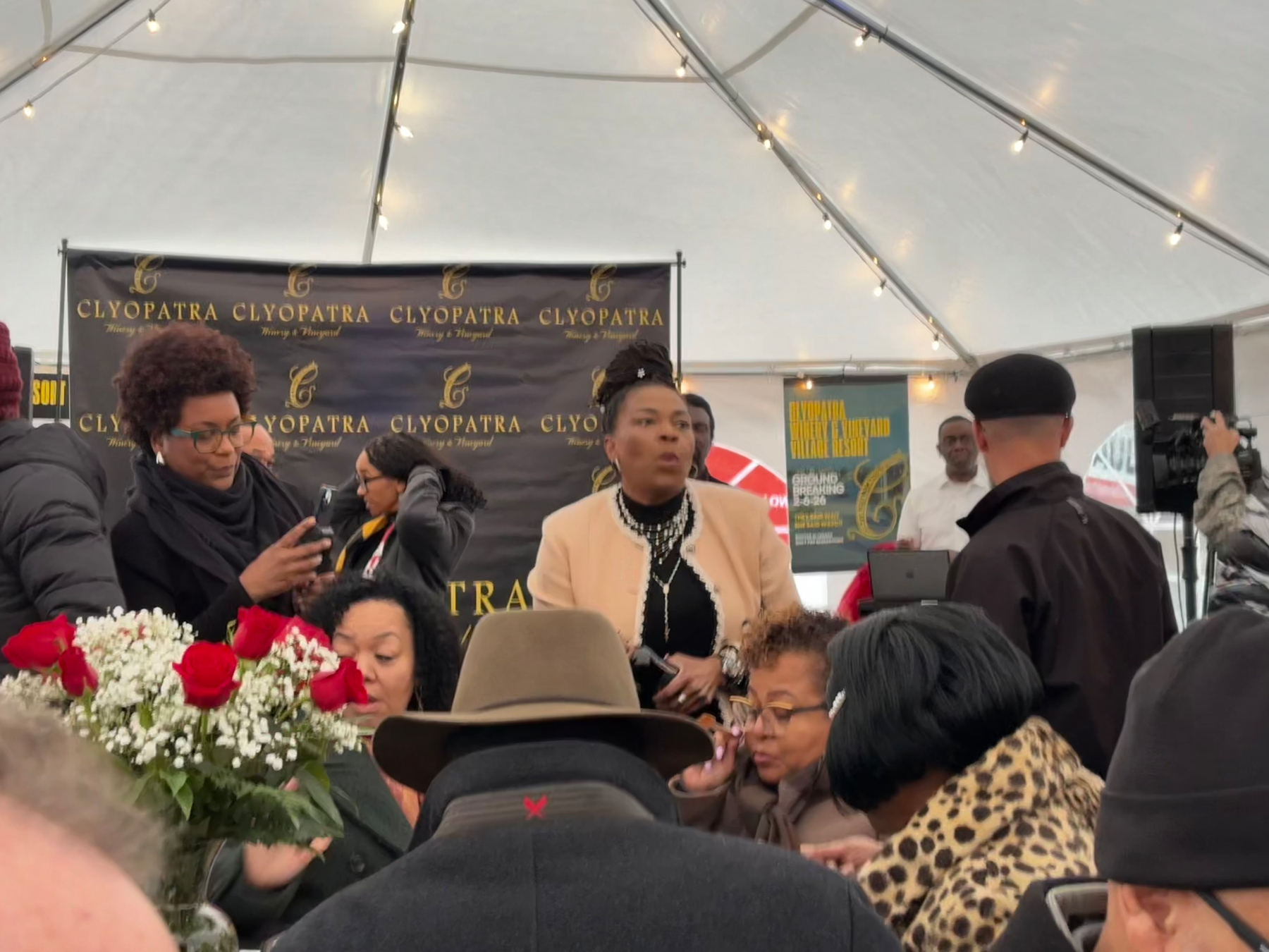 Auto-generated description: A group of people is gathered under a tent with a backdrop that includes the word Cleopatra, along with a bouquet of red roses on a table in the foreground.