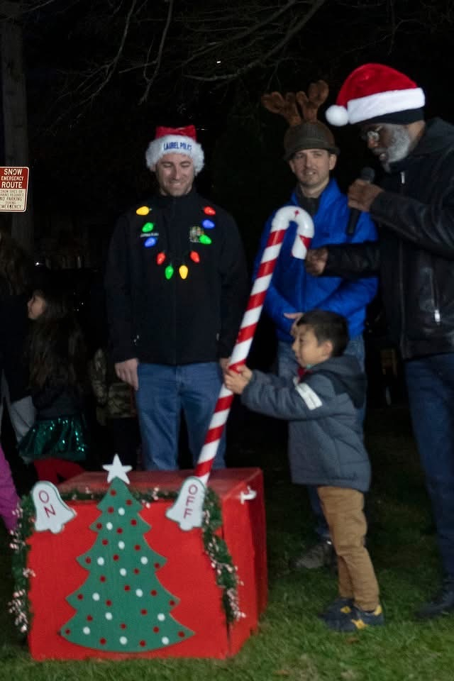 Auto-generated description: A young boy wearing a winter coat pulls on a candy cane lever attached to a red box with a Christmas tree design, surrounded by four adults in festive attire, including Santa hats and string lights.
