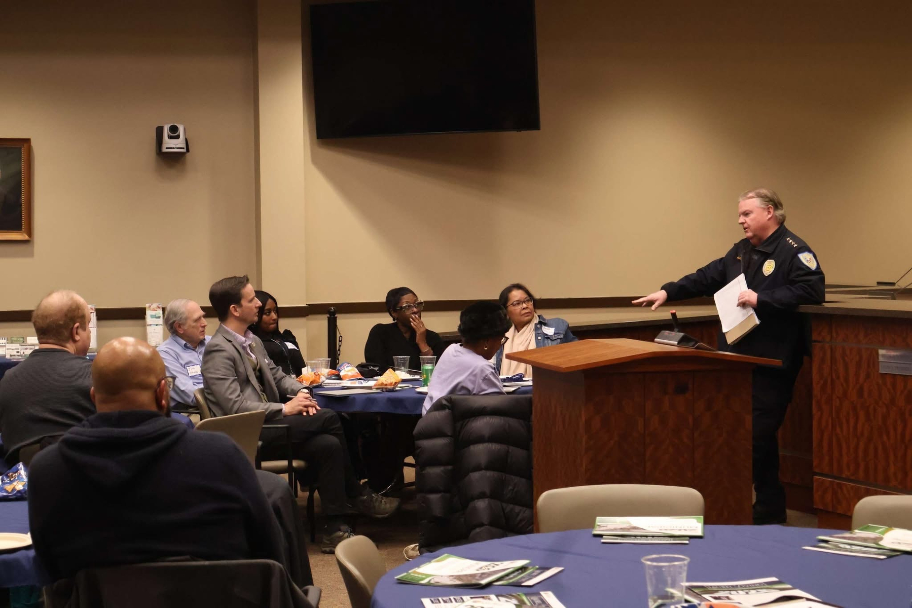Auto-generated description: A group of people sits at tables listening to a speaker, who appears to be a police officer, standing at a podium in a room.