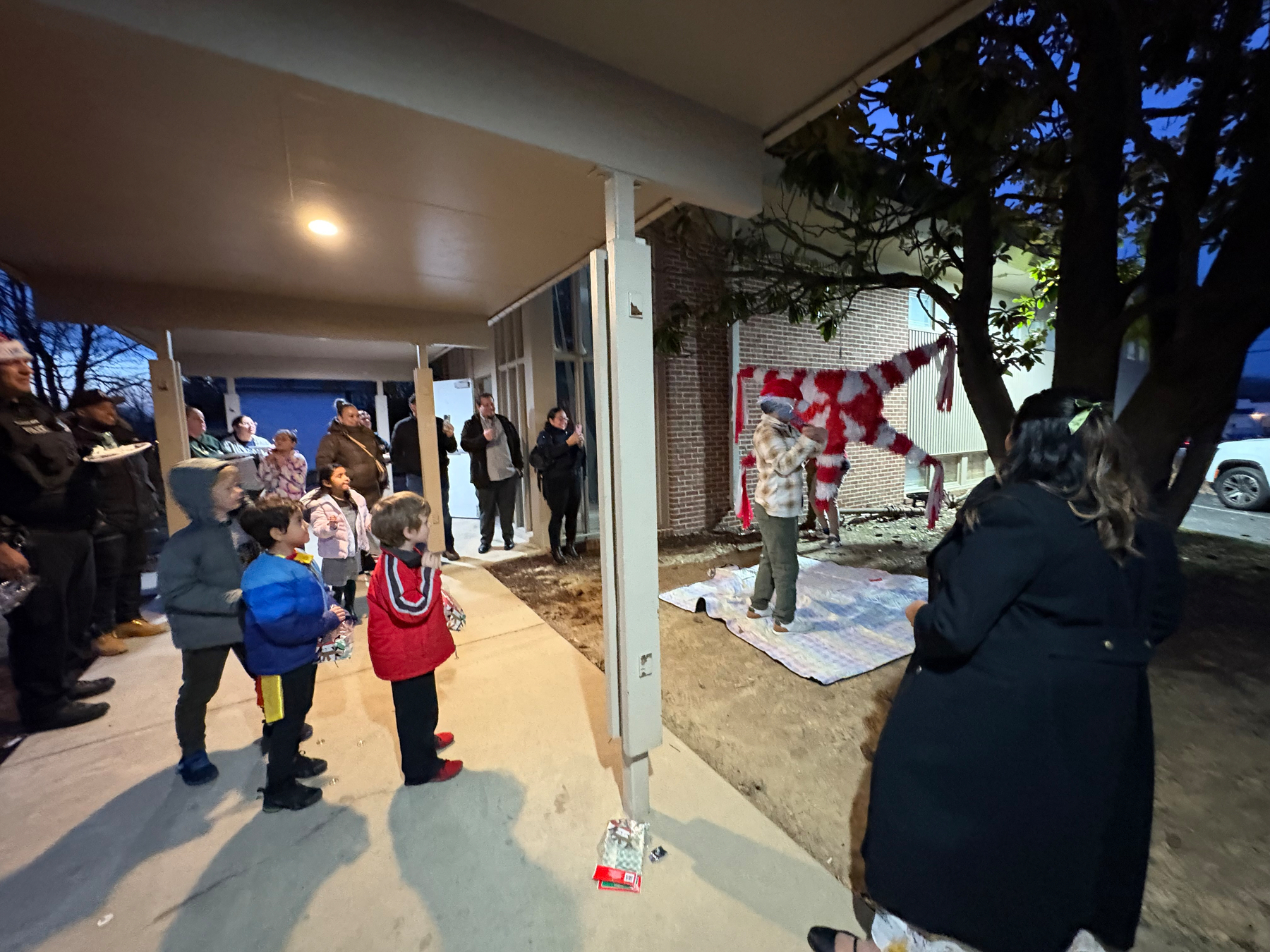 Auto-generated description: A group of people, including children, are gathered outdoors near a building, watching a person in front of a striped hanging decoration.